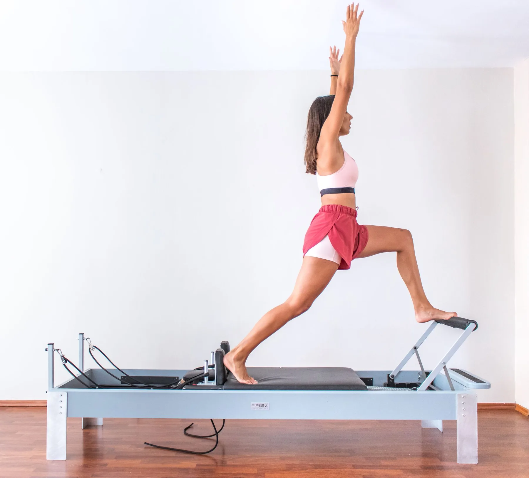 Pratiquante en fente sur un Reformer, bras levé, sur fond blanc