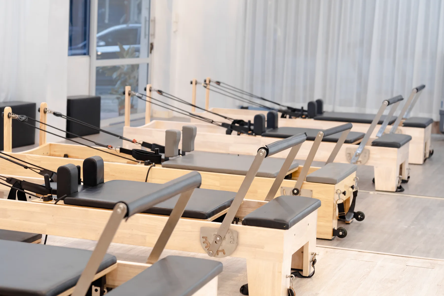 Rangée de Reformers en bois clair dans un studio de Pilates lumineux, voilages blancs et lumière naturelle